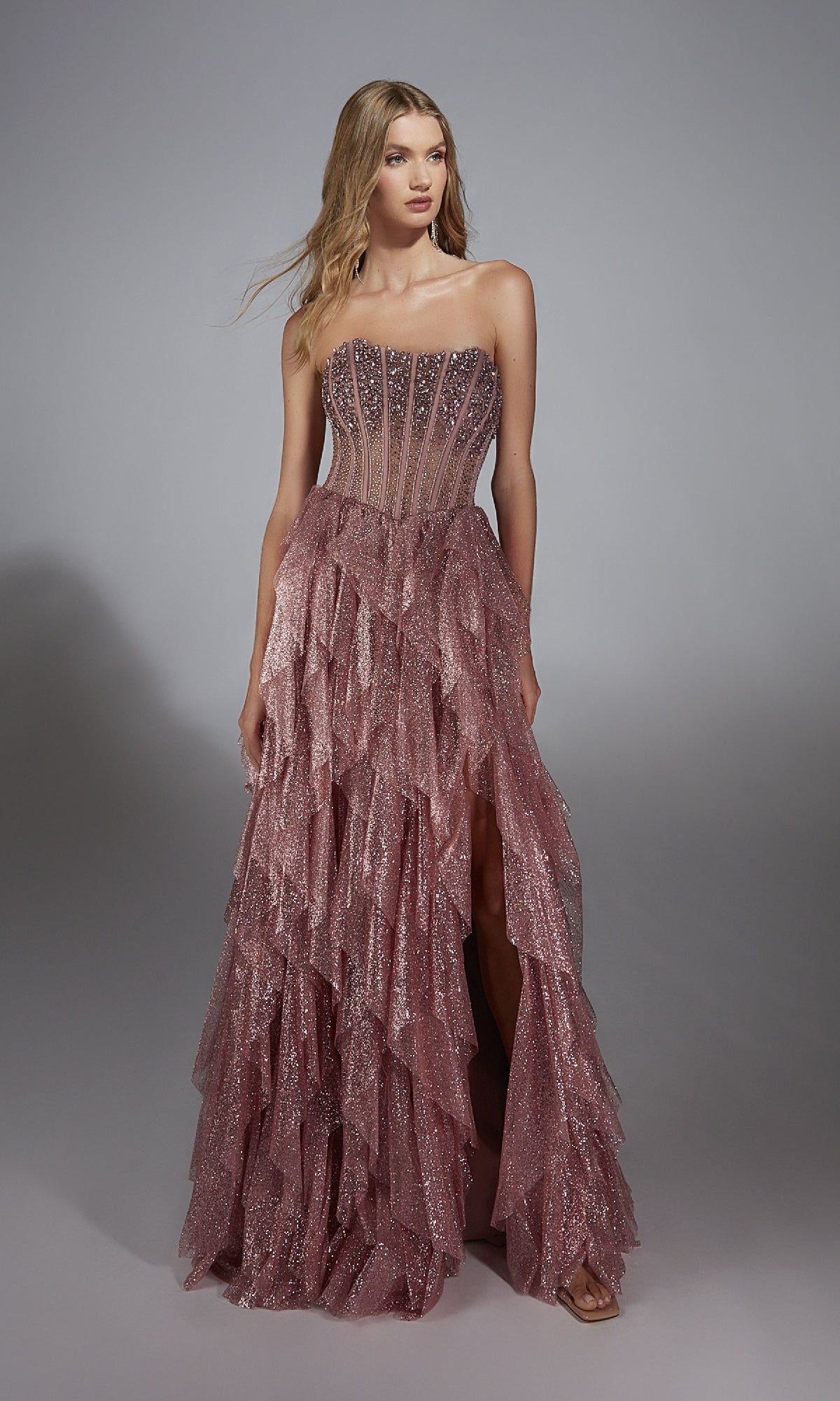 A woman models the Alyce 61762 Strapless Long Glitter A-Prom Dress, featuring a sparkling pink sheer corset bodice and layered ruffled skirt, standing against a plain gray background.